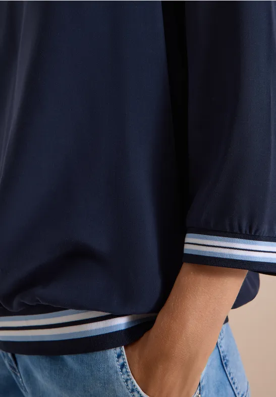 Nahaufnahme einer Person in dunkelblauer Sportjacke mit gestreiften Bündchen und blauer Jeans, Hand in Tasche.