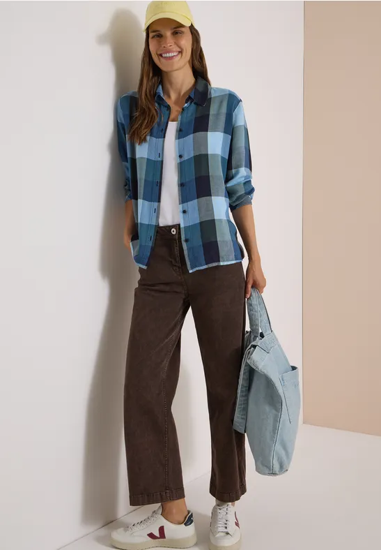 Frau in blau-kariertem Hemd, weißem Shirt, brauner Hose, Sneakern und gelber Cap, hält eine Jeans-Tasche.