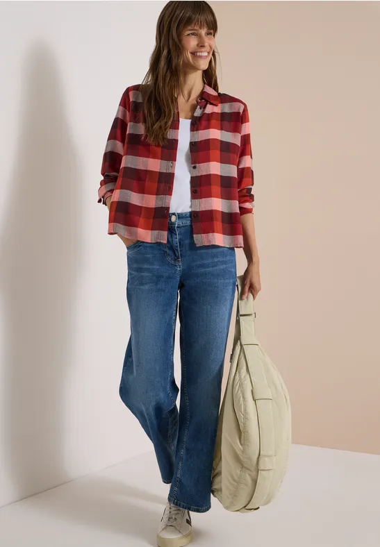 Junge Frau in karierter roter Hemdjacke, weißem Top, blauen Jeans und großer beiger Tasche lächelt.