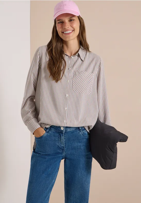 Frau in grau-weiß gestreiftem Hemd, Jeans und rosa Cap, hält eine schwarze Jacke in der Hand.