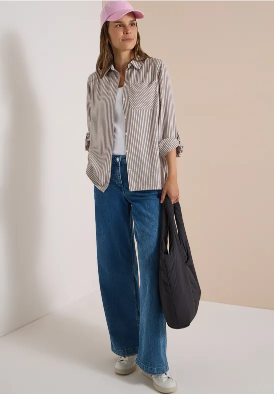 Frau in hellem Overshirt, weißem Shirt und weiten Jeans, mit rosa Cap und schwarzem Beutel auf neutralem Hintergrund.