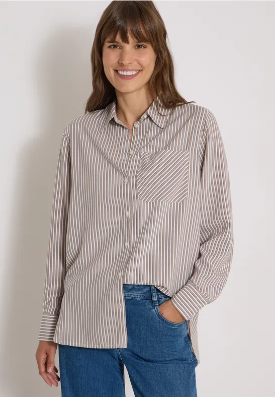 Eine lächelnde Frau trägt eine beige-weiß gestreifte Bluse und blaue Jeans vor hellem Hintergrund.
