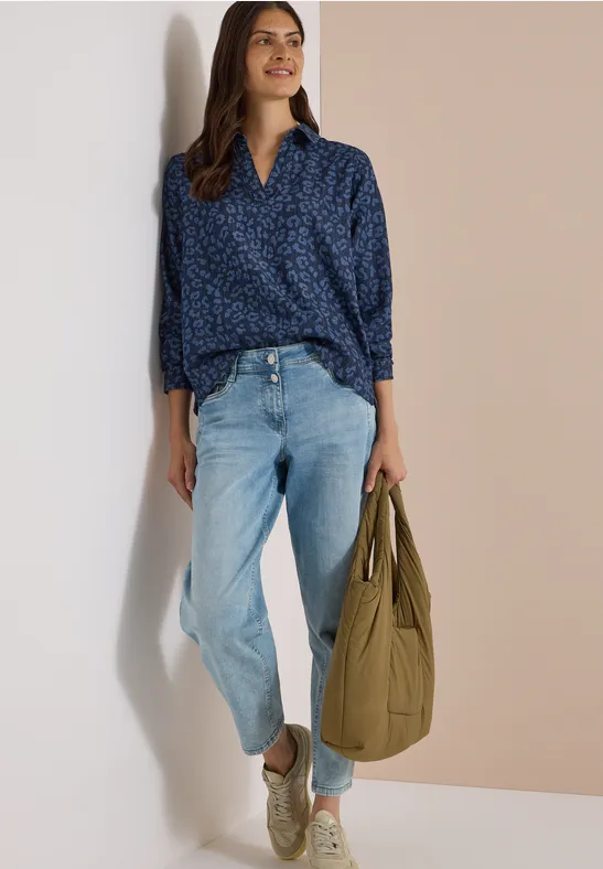 Frau lehnt lässig an einer Wand, trägt eine blaue Bluse, helle Jeans und weiße Sneaker, hält eine grüne Tasche.