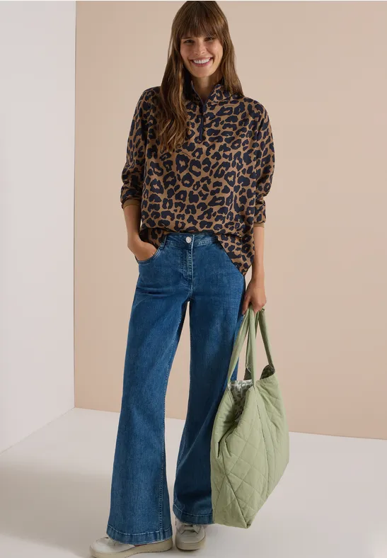 Frau in Leo-Bluse und weitem Jeans, mit großer grüner Tasche, lächelt vor hellem Hintergrund.