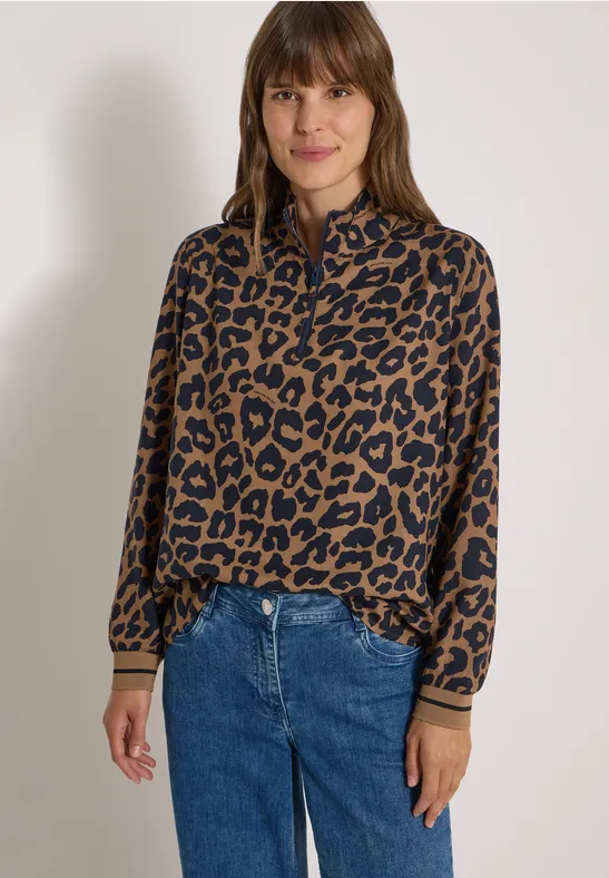 Eine Frau trägt eine braun-schwarze Leopardenmuster-Bluse und blaue Jeans vor hellem Hintergrund.