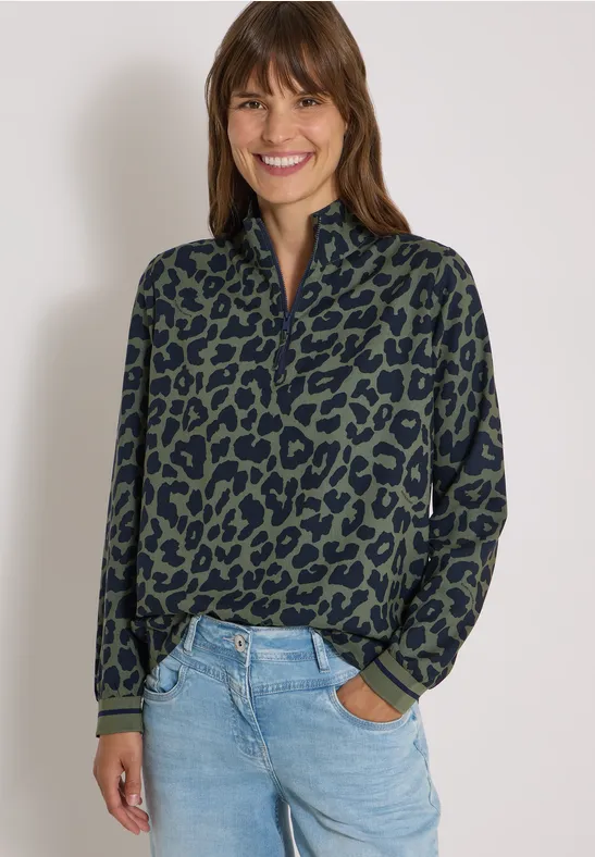 Eine lächelnde Frau trägt einen grünen Leo-Pullover und blaue Jeans vor hellem Hintergrund.