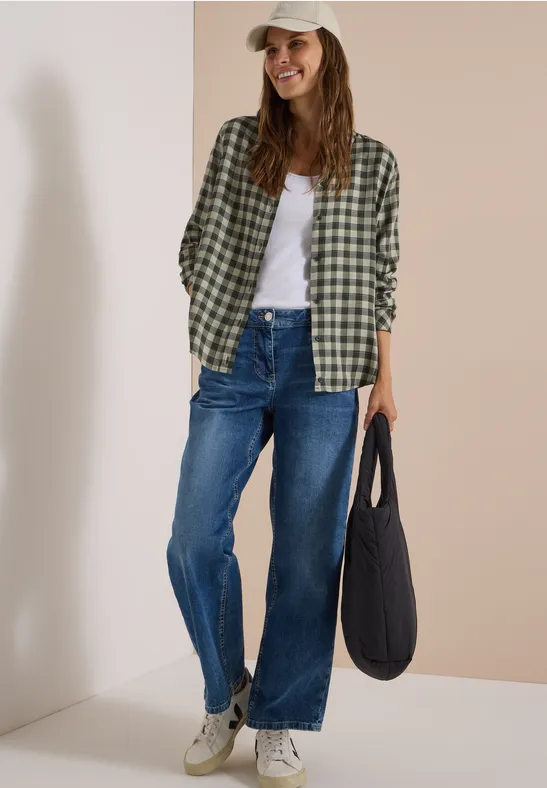 Femme en chemise à carreaux, débardeur blanc, jean large, baskets et casquette, tenant un sac noir, souriant devant un mur beige.