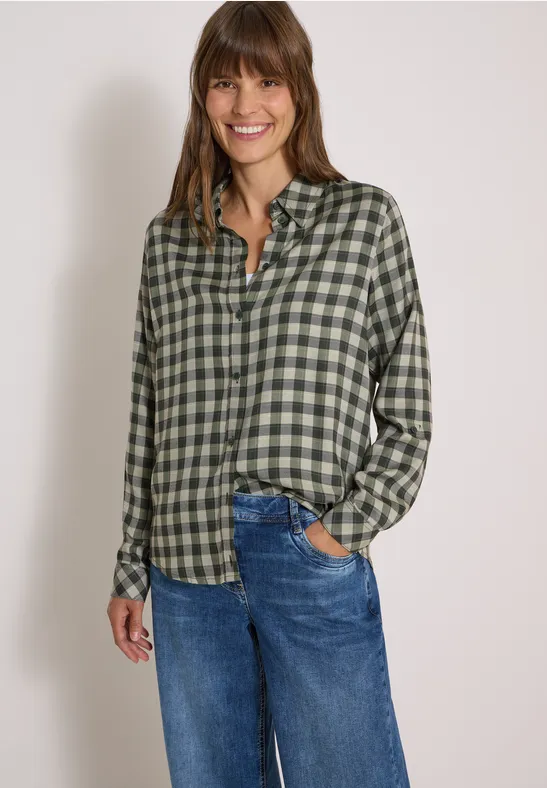 Femme portant une chemise verte à carreaux et un jean bleu, souriante, main droite dans la poche.