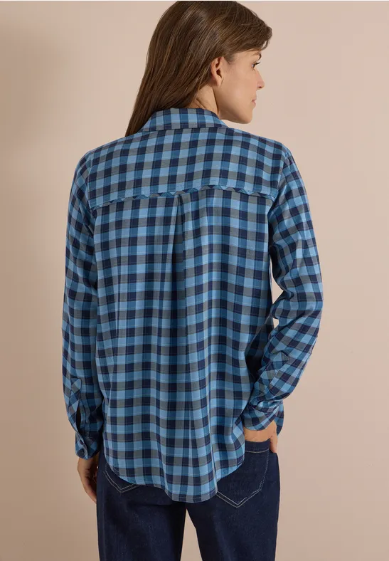 Eine Person von hinten in einem blauen, karierten Hemd vor beigem Hintergrund.