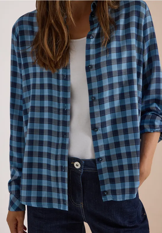 Person trägt blau kariertes Hemd über weißem Top und dunklen Jeans