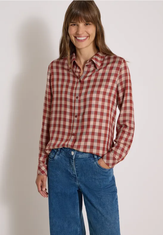 Femme souriante portant une chemise à carreaux et un jean devant un fond neutre.