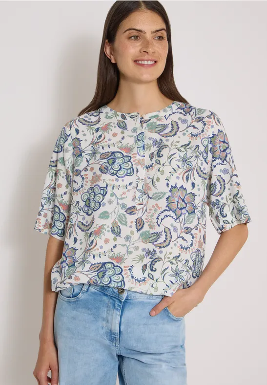 Kobieta w białej bluzce z pastelowym kwiatowym wzorem, luźny krój, w połączeniu z jasnymi jeansami.