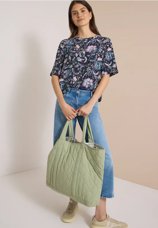 Frau in gemusterter Bluse, Jeans und mit großer gesteppter grüner Tasche.