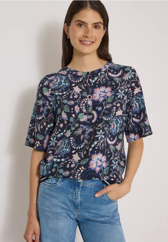 Junge Frau mit dunkler, floraler Bluse und blauen Jeans lächelnd vor hellem Hintergrund.