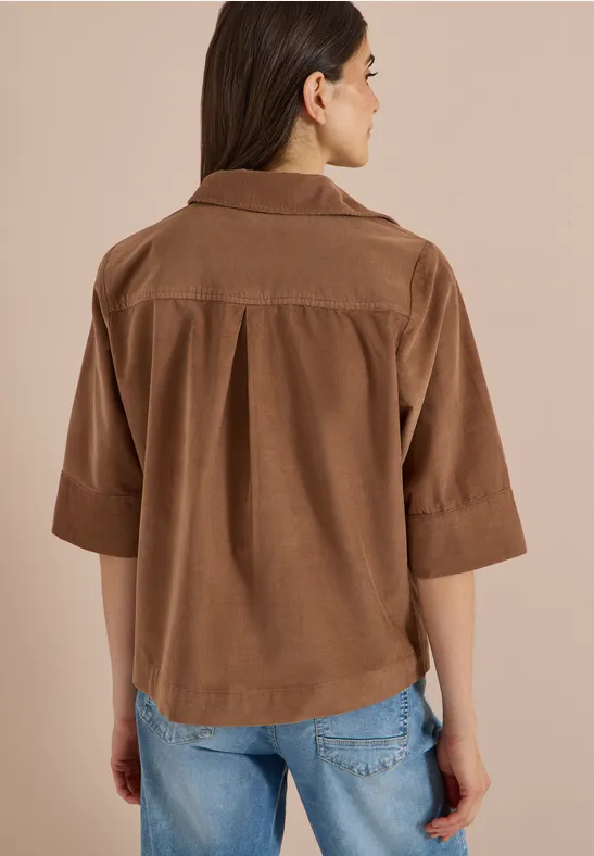 Frau von hinten in lockerer brauner Cord-Hemdbluse mit 3/4-Ärmeln und blauen Jeans vor beigem Hintergrund.