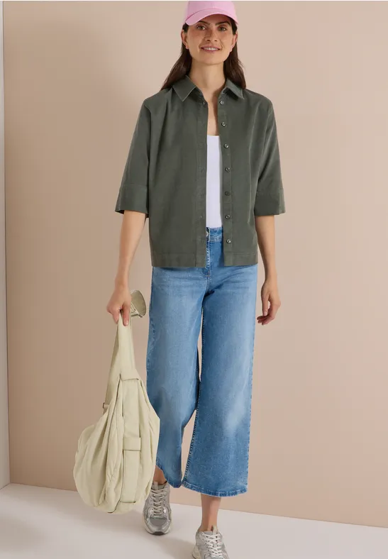 Frau in weitem Jeans-Culotte, olivgrünem Hemd, weißem Top, Sneakern und pinker Kappe trägt Tasche.