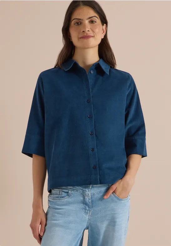 Eine Frau in blauem Hemd mit kurzen Ärmeln und heller Jeans vor neutralem Hintergrund.
