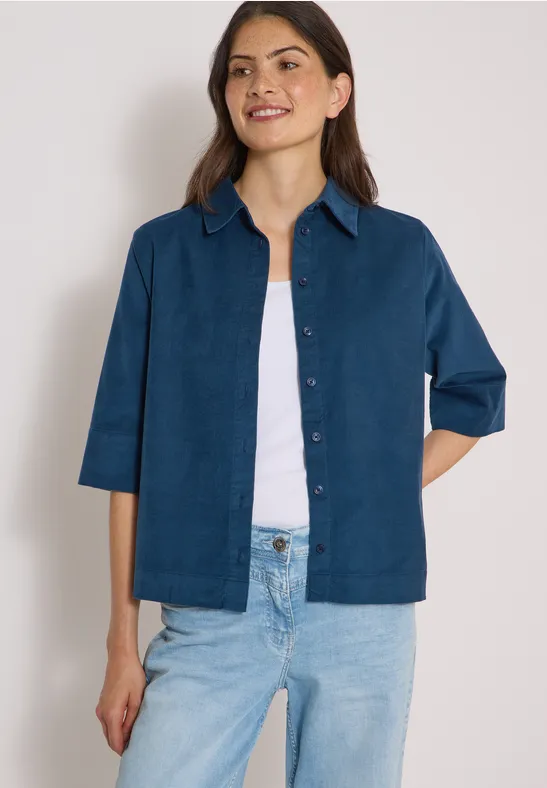 Frau trägt blaues Jackett über weißem Top und Jeans, steht vor neutralem Hintergrund.