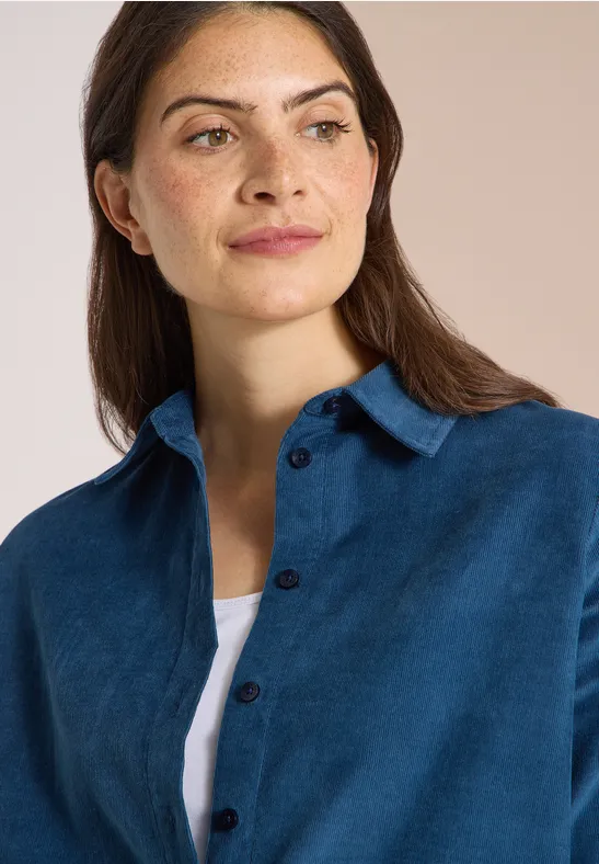 Eine Frau mit braunen Haaren trägt ein blaues Cordhemd über einem weißen Top vor hellem Hintergrund.