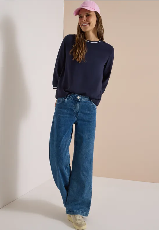 Frau in weitem Jeans, marineblauem Pullover und rosa Cap, lächelnd vor hellem Hintergrund.
