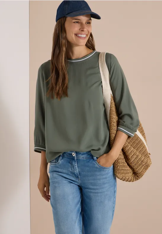 Eine Frau in Jeans, grüner Bluse und Cap trägt lächelnd eine geflochtene Tasche.