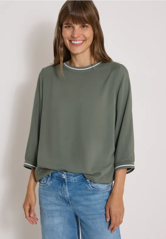 Frau in grüner Bluse mit weißen Bündchen und blauen Jeans lächelt.