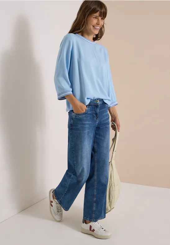 Frau lehnt an Wand, trägt hellblaues Oversize-Shirt, weite Jeans, weiße Sneaker und hält eine Tasche.