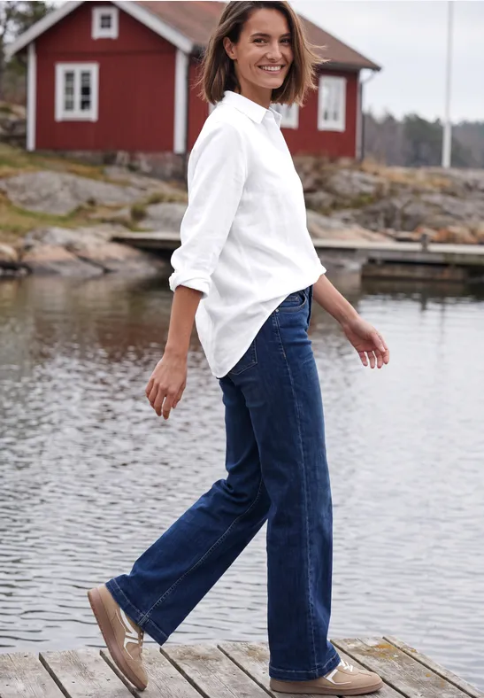 Eine Frau in weißem Hemd und Jeans geht lächelnd auf einem Steg vor einer roten Hütte am Wasser.