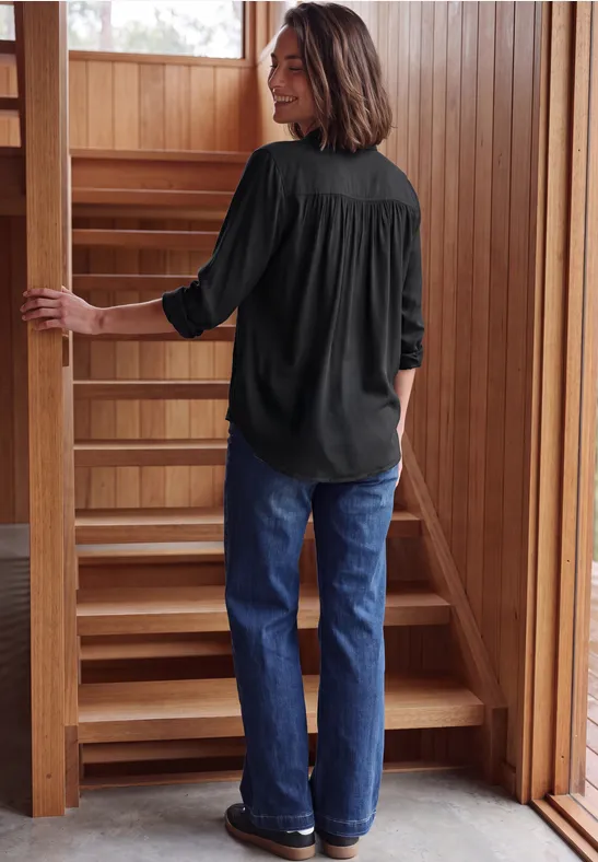 Vrouw in donkere blouse en spijkerbroek loopt een houten trap op in een warme, houten omgeving.