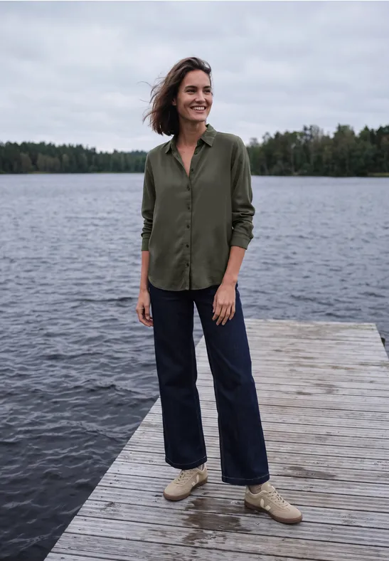Een vrouw staat op een houten steiger aan het meer, gekleed in een groen overhemd, jeans en lichte schoenen.