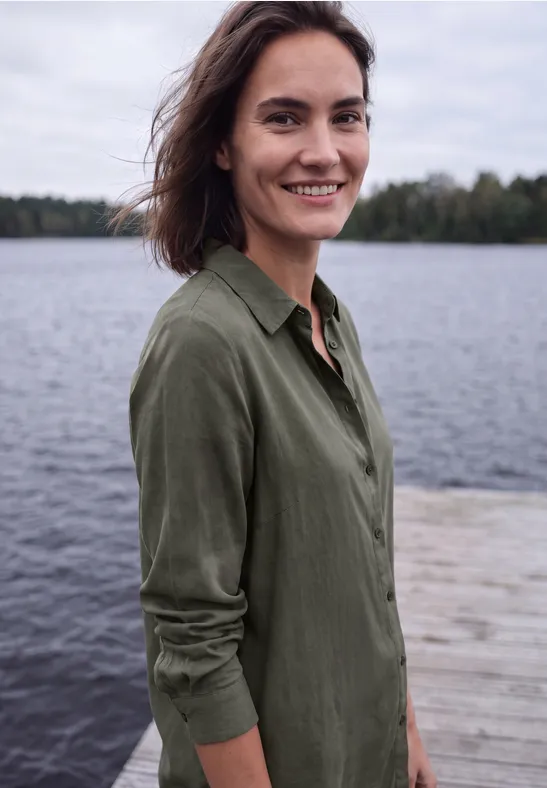 Een glimlachende vrouw staat op een steiger aan een meer, draagt een groen overhemd en kijkt in de camera.