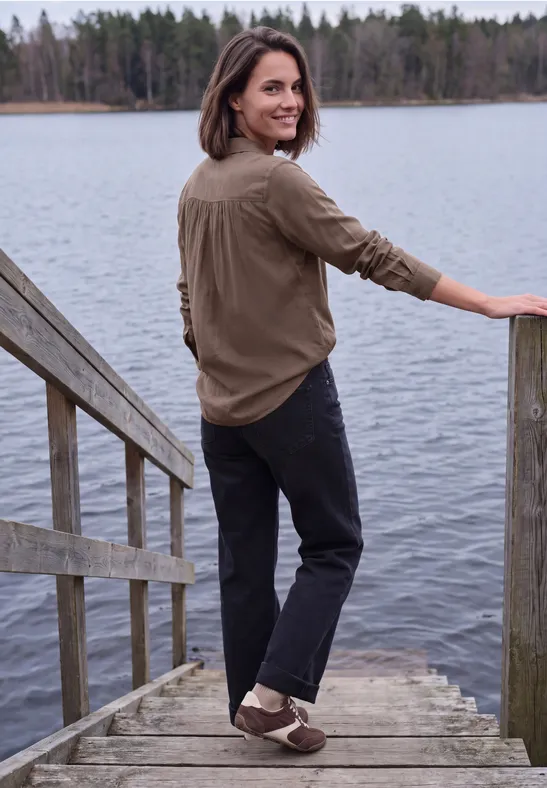 Eine Frau steht lächelnd auf einem Steg an einem See, trägt braunes Hemd und schwarze Hose.