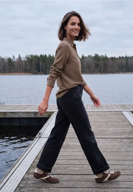 Frau spaziert lächelnd auf einem Steg am See, trägt khakifarbenen Pullover, dunkle Hose und braune Sneaker.