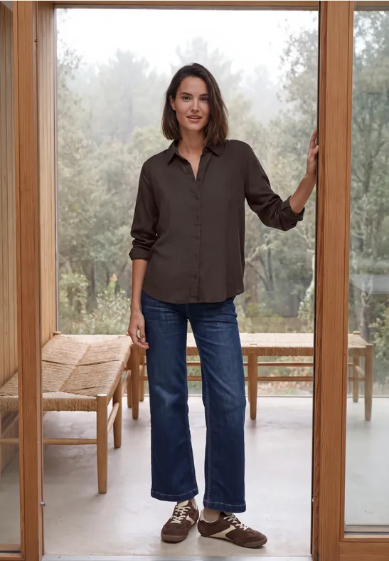 Eine Frau steht lässig in einer Türöffnung, trägt braunes Hemd, gerade geschnittene Jeans und braune Schuhe.