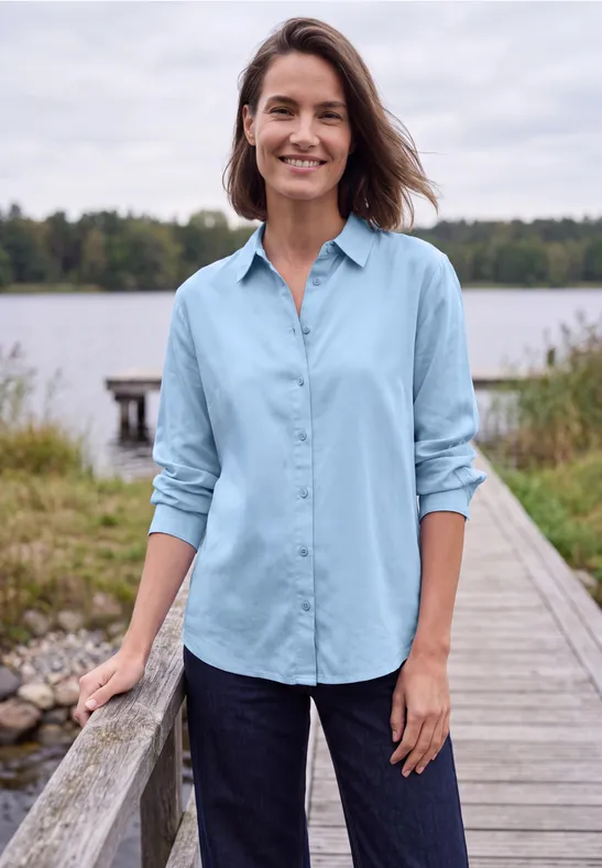 Vrouw in blauw overhemd, lachend op een steiger bij een meer, omringd door gras en bomen.