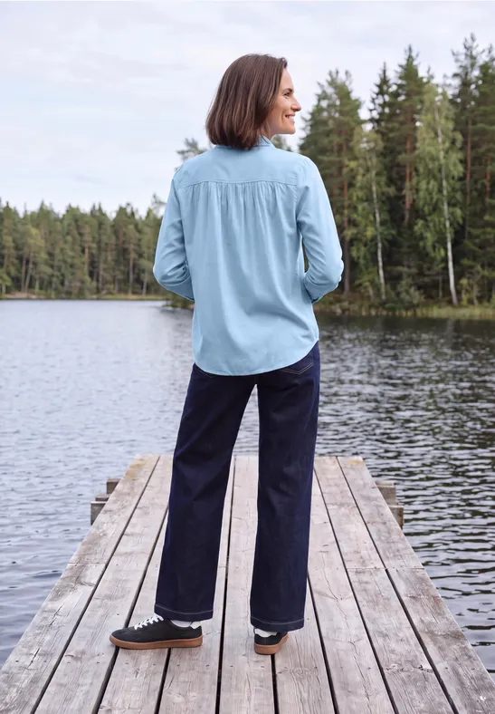 Een vrouw in lichtblauw overhemd en donkere broek staat op een steiger aan een meer, omringd door bos.