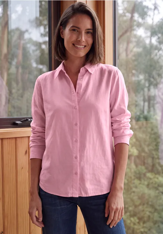 Eine Frau in rosa Hemd und Jeans lächelt vor einer Fensterkulisse mit Bäumen.