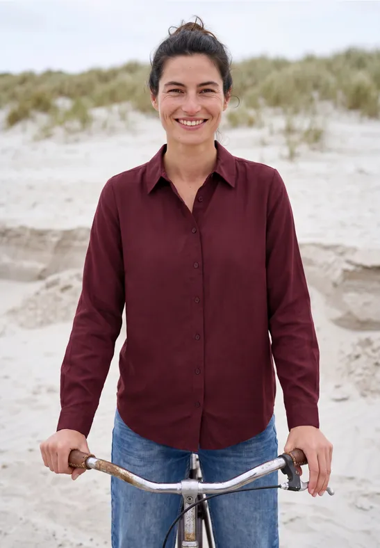 Eine lächelnde Frau mit rotem Hemd und Jeans steht am Strand und hält ihr Fahrrad.