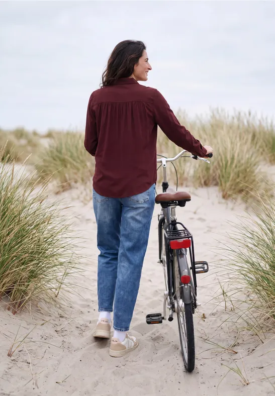 Eine Person schiebt ihr Fahrrad durch sandige Dünen, umgeben von Strandgras bei bewölktem Himmel.