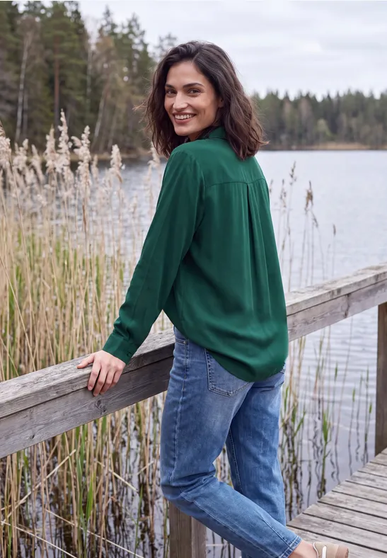 Vrouw in groen overhemd lacht op houten steiger aan een meer, omringd door riet en bos op de achtergrond.