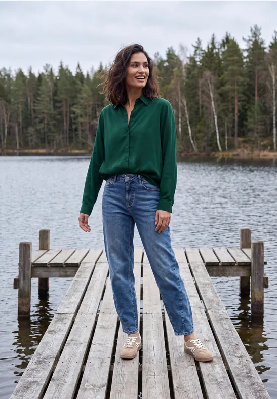 Vrouw in groen shirt, spijkerbroek en lichte schoenen staat op een houten steiger bij een meer, bos op achtergrond.