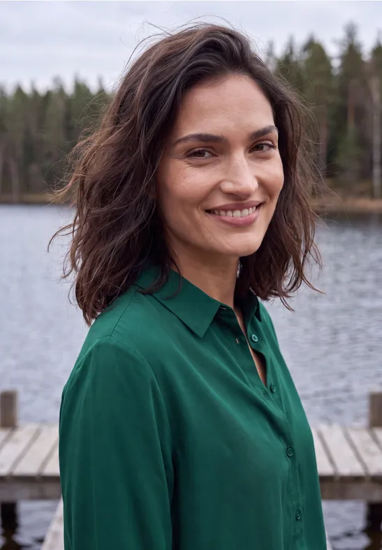 Een lachende vrouw in een groen blouse staat op een steiger bij een meer met bos op de achtergrond.