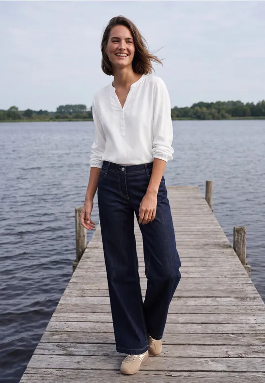 Eine lächelnde Frau in weißer Bluse und dunkler Jeans steht auf einem Steg am See.