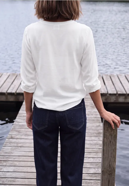 Rückansicht einer Frau mit weißem Oberteil und Jeans auf einem Holzsteg am Wasser