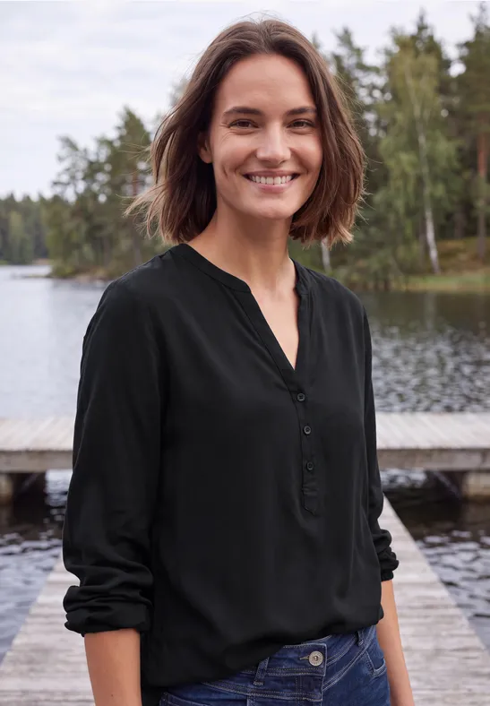 Een glimlachende vrouw in een zwarte blouse staat op een steiger aan een meer met bomen.