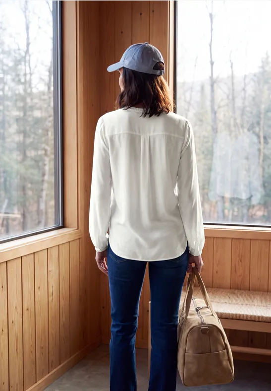 Frau trägt Baseballcap, weiße Bluse und Jeans, steht mit Tasche vor großem Fenster in Holzraum.