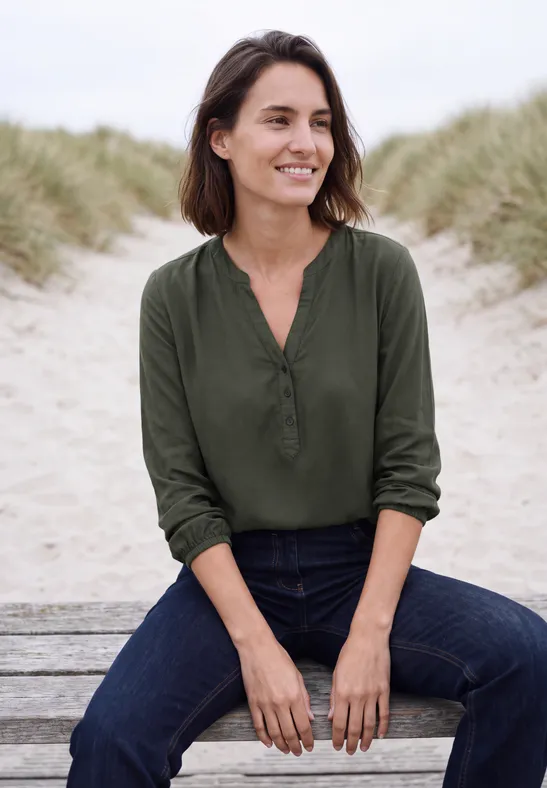 Een lachende vrouw in een groene blouse en jeans zit ontspannen op een bank bij de zandduinen.