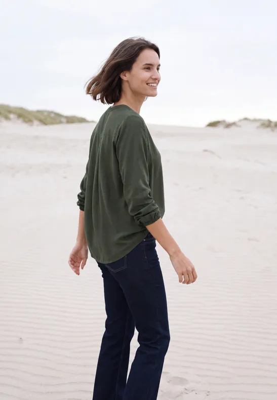 Een vrouw in groene trui en spijkerbroek wandelt over het strand.