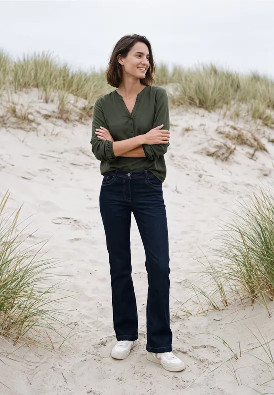 Vrouw in groen shirt en spijkerbroek, glimlachend met armen over elkaar op zandduinen.