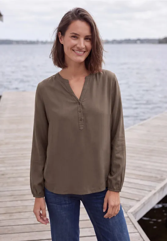 Een glimlachende vrouw staat op een steiger bij het water, met een olijfgroen blouseje en blauwe jeans.
